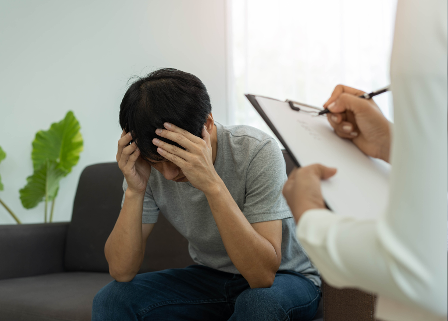 A person sitting on a couch with their head in their hands and another person holding a clipboard in front of them